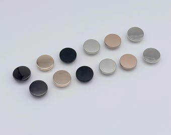 May include: A collection of round, metallic buttons in various colors, including black, silver, and rose gold. The buttons are arranged on a white surface, showcasing their smooth, reflective surfaces. These buttons are suitable for sewing and craft projects.