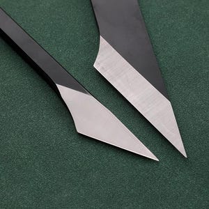 Puede incluir: Dos cuchillos artesanales negros y plateados con hojas anguladas. Las hojas tienen un acabado plateado y los mangos son negros. Los cuchillos están sobre un fondo verde oscuro.