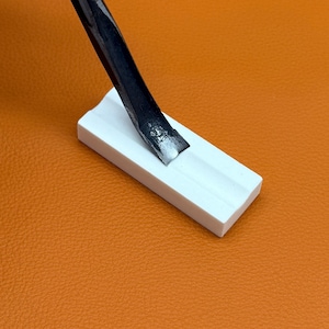 May include: A white rectangular block with a central groove, featuring a metallic chisel tip resting within the channel. The object is positioned against a textured orange surface, highlighting the contrast between the white material and metal.