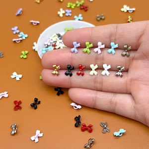 May include: A collection of small, colorful bow-shaped embellishments. The bows are in various colors, including gold, black, red, white, and pastel shades of pink, green, and blue. Some bows are held in a hand, while others are scattered on a brown surface.
