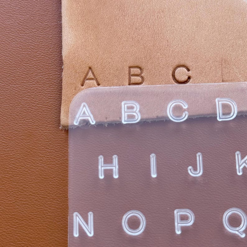 May include: Close-up of leather crafting tools. The image shows the letters A, B, and C embossed on tan leather. A clear plastic stencil with the letters A, B, C, D, H, I, J, K, N, O, P, and Q is also visible.