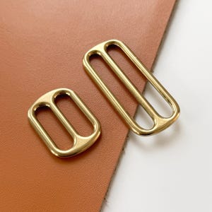 Op de afbeelding: Twee goudkleurige metalen gespen. E&eacute;n is rechthoekig met twee staven, de andere is kleiner met een vergelijkbaar ontwerp. De gespen liggen op een bruin leren oppervlak.