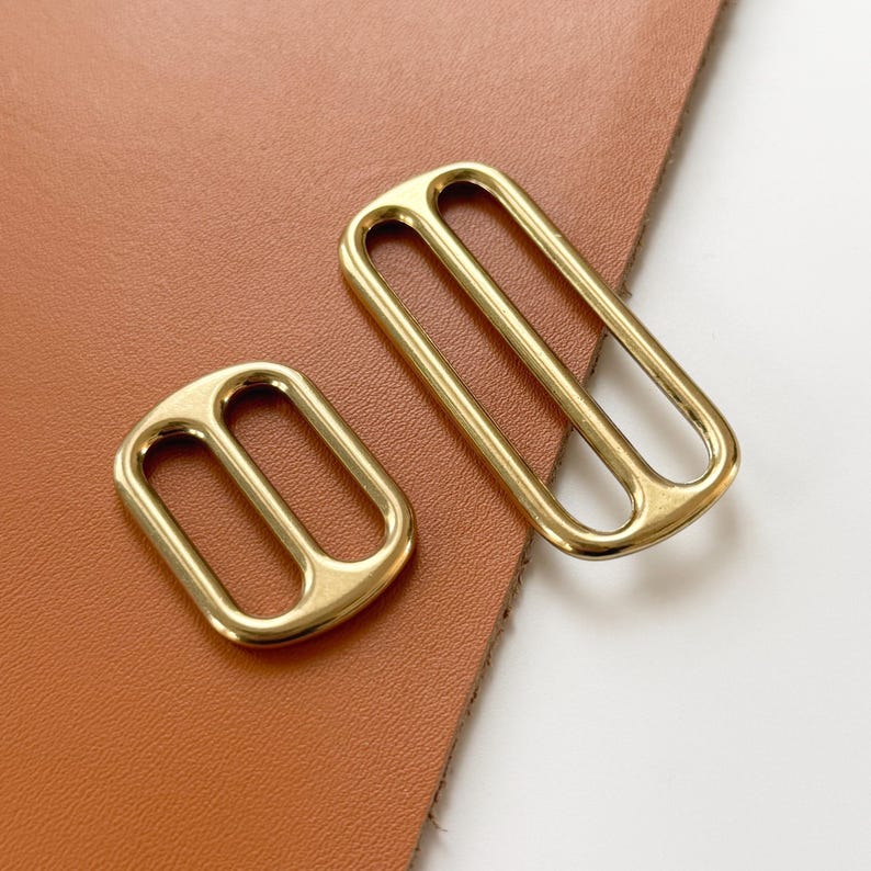 Op de afbeelding: Twee goudkleurige metalen gespen. E&eacute;n is rechthoekig met twee staven, de andere is kleiner met een vergelijkbaar ontwerp. De gespen liggen op een bruin leren oppervlak.