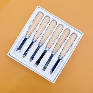 May include: A set of six wood carving chisels with light-colored wooden handles and silver metal blades, arranged in a white plastic tray. Each tool features a distinct blade shape, including straight, angled, and gouge tips for woodworking.