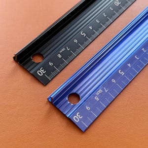 Puede incluir: Dos reglas de metal, una negra y otra azul, cada una con una escala de 30 cm. Las reglas tienen un diseño ranurado y un agujero en un extremo. Los números y las marcas son blancos.