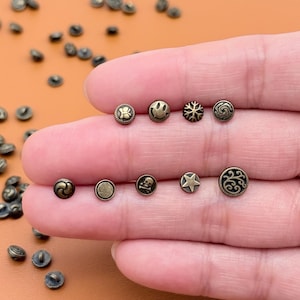 以下が含まれることがあります： 花、スマイリーフェイス、雪の結晶、渦巻き、星など、さまざまなデザインの小さなアンティーク真鍮色のボタンのコレクション。ボタンは丸く、わずかに隆起したデザインです。一部のボタンには、ドクロのデザインがあります。