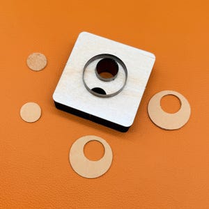 May include: A square wooden die cutter featuring two concentric circular metal blades. The tool is accompanied by two small circular leather cutouts and one tiny circular cork cutout, all arranged on a flat orange surface.