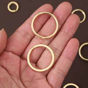 May include: Close-up of several gold-colored metal rings. The rings are circular with a small opening. The rings are held in a person's hand against a brown background. The rings are likely used for jewelry making or crafting.