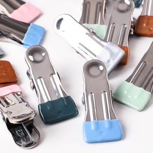 May include: A selection of metal binder clips with coloured rubber ends. The clips have a shiny silver finish and are accented with blue, pink, green, and brown rubber. These clips are suitable for organising papers and other items.