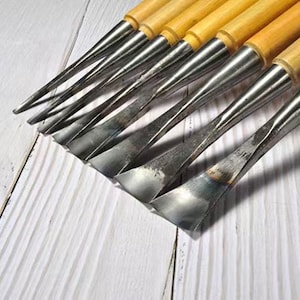 Può includere: Un set di utensili per intaglio del legno con manici in legno e lame in metallo. Le lame hanno varie forme e dimensioni e sono disposte su una superficie di legno bianco. Progettati per lavori di falegnameria dettagliati.