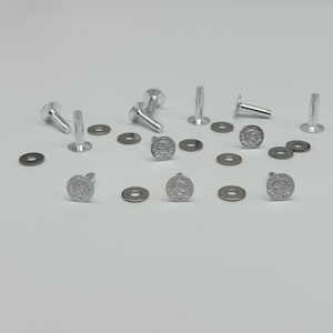 Könnte beinhalten: Eine Sammlung silberfarbener Metallnieten und passender flacher, runder Unterlegscheiben. Jede Niete hat einen runden Kopf mit einem geprägten Sonnenstrahlenmuster. Die Beschlagteile sind verstreut und zeigen die Stifte aus verschiedenen Winkeln.