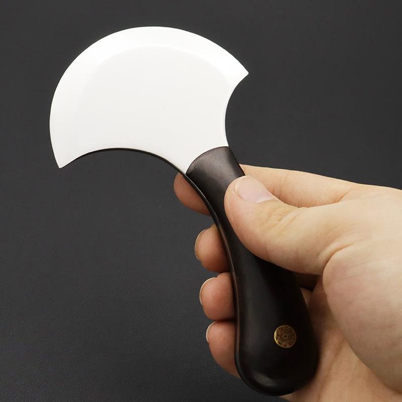 Op de afbeelding: Een leerambachtmes met een gebogen, wit lemmet en een donkerbruin handvat. Het handvat heeft een klein, goudkleurig embleem bij de basis. Het mes wordt in een hand gehouden tegen een donkere achtergrond.
