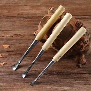 Könnte beinhalten: Drei Holzschnitzmesser mit hellfarbenen Holzgriffen und silberfarbenen Klingen. Die Werkzeuge sind diagonal auf einer Holzoberfläche angeordnet, mit einer geschnitzten Holzskulptur im Hintergrund. Die Stecheisen sind für detaillierte Holzarbeiten konzipiert.