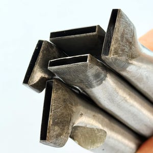 Puede incluir: Primer plano de varios punzones metálicos con aberturas rectangulares. Las herramientas son de metal color plata y tienen un aspecto desgastado y texturizado. Los punzones están dispuestos de forma ligeramente superpuesta.