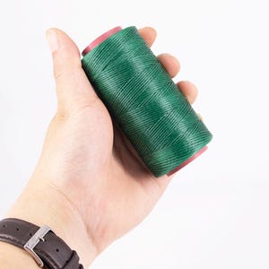 Può includere: Una mano che tiene una bobina di filo verde scuro, con un orologio marrone al polso. Il filo è avvolto attorno a un'anima rosa. Lo sfondo è bianco.