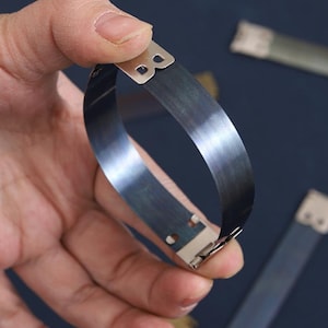 Puede incluir: Una pulsera de metal gris oscuro curvada con cierres plateados. La pulsera se sostiene en una mano, con otras piezas similares visibles en el fondo. El metal tiene una superficie reflectante.