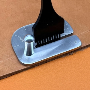 Peut inclure: Un gabarit d'angle en métal argenté pour le travail du cuir avec une poignée, portant les marquages R20 et R5. Un poinçon de couture noir à plusieurs dents est inséré dans la fente de guidage, posé sur un cuir beige avec des coutures.