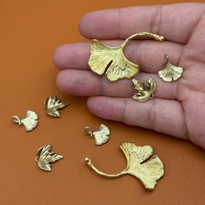 Könnte beinhalten: Eine Sammlung goldfarbener Anhänger und Anhänger in Blattform. Die Anhänger haben Ginkgo- und Ahornblatt-Designs in verschiedenen Größen. Das Metall hat eine strukturierte Oberfläche, und einige Teile haben kleine Schlaufen zum Befestigen an Schmuck.