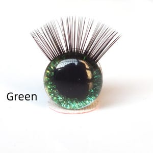 May include: A green glass eye with black eyelashes. The eye has a dark pupil and a glittery green iris. The word "Green" is printed in black text below the eye.