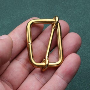 May include: A gold-colored metal buckle with a rectangular frame and a triangular piece attached. The buckle is held in a hand against a green background. The metal has a shiny finish.