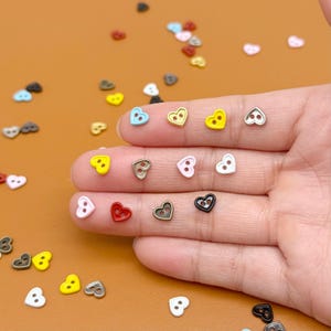 May include: A selection of small, heart-shaped buttons in a variety of colours, including yellow, pink, blue, red, and black. The buttons have two holes for sewing and are scattered on a brown surface. Some buttons are held in a hand.