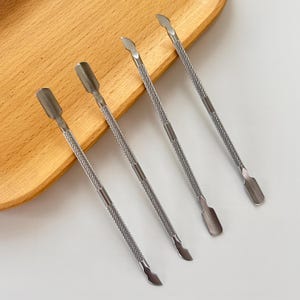 May include: Four stainless steel cuticle tools with textured handles and various tip shapes, including a scoop, a curved edge, and a pointed end. The tools are arranged on a light brown wooden tray.