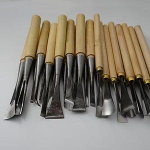Könnte beinhalten: Eine Sammlung von Holzbearbeitungswerkzeugen mit Holzgriffen und Metallklingen. Die Klingen haben verschiedene Formen, darunter flach, gebogen und abgewinkelt, für detaillierte Holzbearbeitungsprojekte.