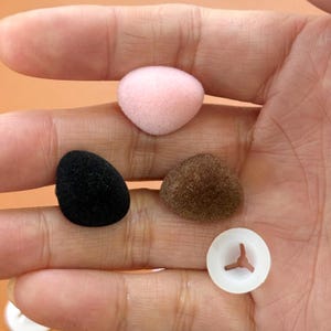 Puede incluir: Una colección de pequeñas fundas de espuma para auriculares de colores y una pieza de plástico blanco con un diseño de tres puntas. Las fundas de los auriculares son de color rosa, negro y marrón. Los artículos se sostienen en una mano.