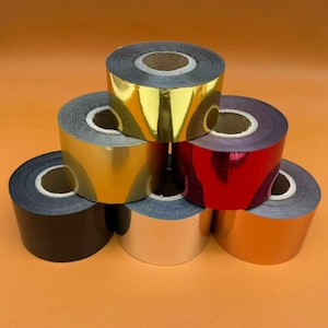 May include: A collection of six rolls of metallic foil arranged in a pyramid shape. The rolls feature reflective surfaces in gold, red, silver, orange, and black, each wound around a central cardboard core, showcasing a smooth and glossy finish.