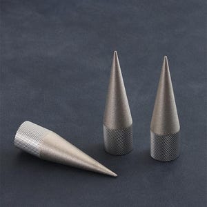 Puede incluir: Tres objetos metálicos en forma de cono, con bases texturizadas y puntas afiladas. Dos están de pie, mientras que el tercero está tumbado de lado. Los objetos son de color plateado y están sobre un fondo oscuro y texturizado.