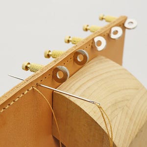 Puede incluir: Primer plano de herramientas de artesanía en cuero. Una pieza de cuero marrón claro se cose a mano con aguja e hilo. Tornillos de latón y arandelas plateadas están adheridos al cuero. El cuero descansa sobre una forma de madera.