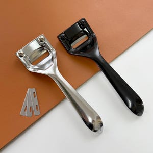 May include: Two leathercraft tools: a silver-colored and a black edge beveler, each with a handle and a blade slot. Three spare blades are also visible. The tools are designed for leatherworking and are set against a brown background.