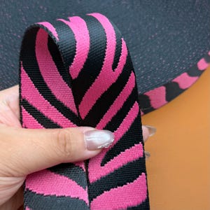 May include: A roll of woven webbing tape featuring a repeating zebra-like pattern in vibrant hot pink and black. The fabric has a distinct ribbed texture and is shown as a continuous strip, highlighting the bold, high-contrast color scheme.