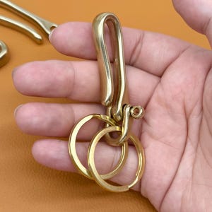May include: A brass key holder with a large hook and two key rings. The key holder is made of shiny, polished brass and is designed to attach to a belt loop or bag. The key rings are round and can hold multiple keys.
