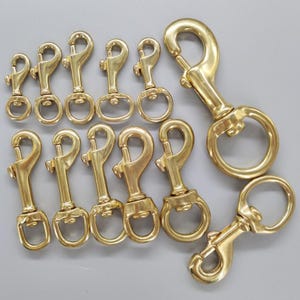 May include: A collection of twelve brass swivel bolt snaps arranged in two rows. Each fastener features a spring-loaded gate and a rotating eyelet at the base. The hardware varies in size, with one significantly larger clip positioned on the right.