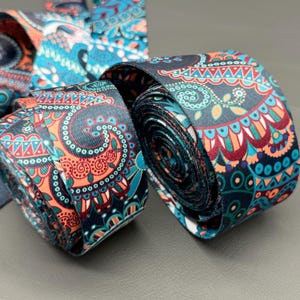 May include: Close-up of two rolled decorative ribbons. The ribbons feature a dark background with a repeating pattern of orange, teal, and blue paisley-like designs. The ribbons are tightly rolled, showcasing the intricate details of the pattern.