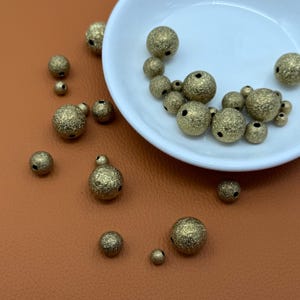 May include: A collection of gold-coloured, textured beads in a white bowl and scattered on a brown surface. The beads vary in size and have a rough, glittery texture with small holes. Suitable for jewellery making or craft projects.
