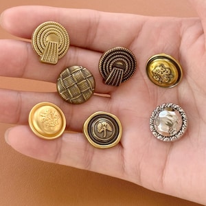 Może przedstawiać: Kolekcja metalowych guzików w stylu vintage w różnych wzorach. Guziki charakteryzują się skomplikowanymi detalami, w tym wzorami tkanymi, teksturami przypominającymi liny oraz wytłoczonymi motywami kwiatowymi i botanicznymi. Paleta kolorów obejmuje odcienie złota, brązu i srebra.