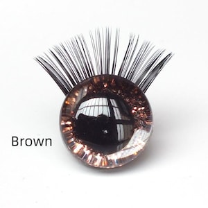 May include: A brown glass eye with black eyelashes. The eye has a reflective surface and is surrounded by a clear, glittery border. The word "Brown" is printed in black text.