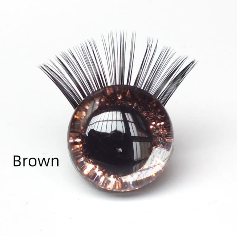 May include: A brown glass eye with black eyelashes. The eye has a reflective surface and is surrounded by a clear, glittery border. The word "Brown" is printed in black text.