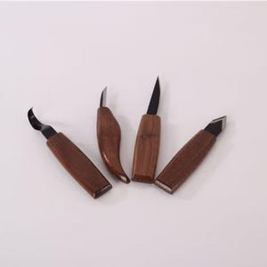 Puede incluir: Cuatro cuchillos para tallar madera con hojas oscuras y mangos de madera. Las hojas tienen diferentes formas, incluyendo un diseño curvo, puntiagudo y en ángulo. Los mangos son de color marrón intenso.