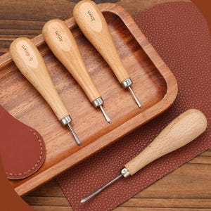 May include: A set of four leather working tools with wooden handles and metal tips. Three tools are arranged in a wooden tray, with the measurements 3mm, 4mm, and 5-6mm etched on the handles. A fourth tool rests on a brown leather surface.
