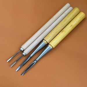 May include: Four wood carving chisels with long cylindrical handles. Two handles are light-colored wood, and two are a darker yellow-toned wood. Each tool features a metal ferrule connecting the handle to a sharp, narrow steel blade tip.