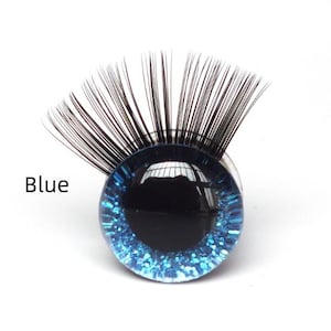 May include: A single, round, blue glass eye with black eyelashes. The eye has a dark center and a sparkling blue iris. The word "Blue" is printed in black text to the left of the eye. This item is likely for crafting or doll making.