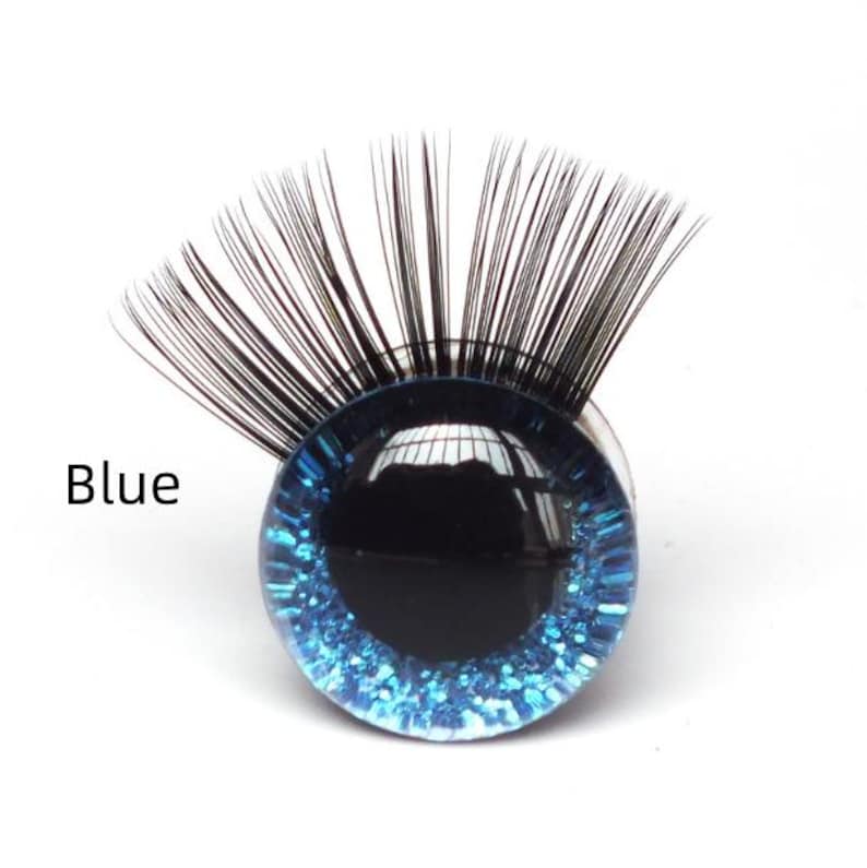 May include: A single, round, blue glass eye with black eyelashes. The eye has a dark center and a sparkling blue iris. The word "Blue" is printed in black text to the left of the eye. This item is likely for crafting or doll making.