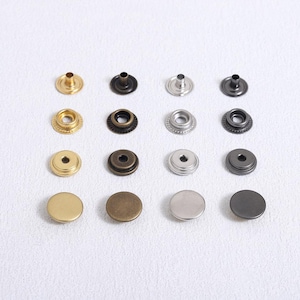 May include: An assortment of metal snap fasteners in various colors, including gold, bronze, silver, and black. The snaps are arranged in rows and columns, showcasing different styles and finishes. The image is well-lit, highlighting the details of each fastener.