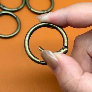 May include: A round, antique brass-coloured metal spring gate ring with a hinged opening mechanism. The circular hardware features a distressed, metallic finish and is designed to function as a connector for bags, keychains, or various craft projects.