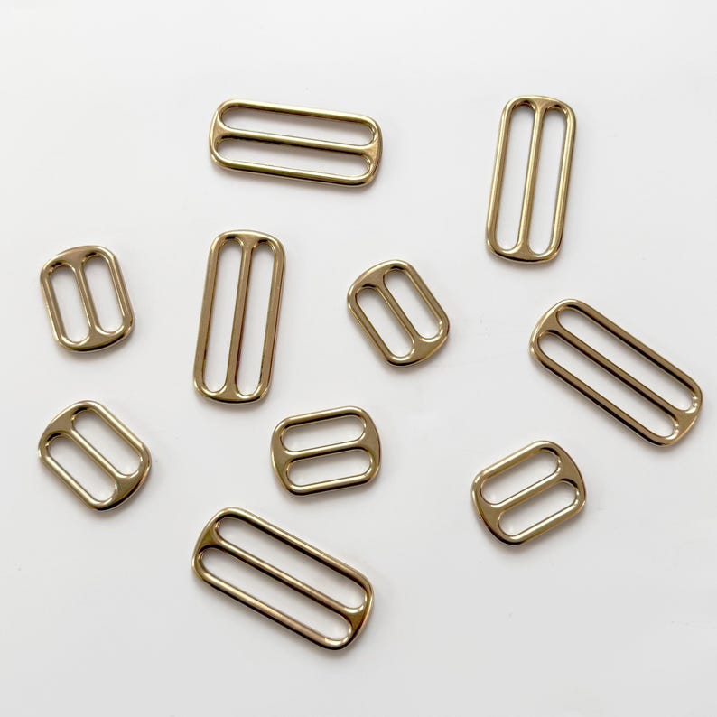 Op de afbeelding: Een verzameling goudkleurige metalen schuifregelaars in verschillende vormen en maten. Deze rechthoekige en ovale schuifregelaars hebben twee parallelle staven en worden gebruikt voor het verstellen van bandjes op kleding of accessoires. De schuifregelaars zijn verspreid over een witte ondergrond.