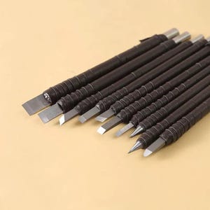 May include: A set of woodworking chisels with dark brown leather-wrapped handles and silver metal blades. The chisels are arranged diagonally, showcasing various blade shapes for detailed carving and shaping of wood.