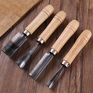 May include: Four wood carving chisels with light brown wooden handles and silver metal collars. The blades are dark gray and vary in shape, with some being flat and others curved. The tools are arranged on a wooden surface.